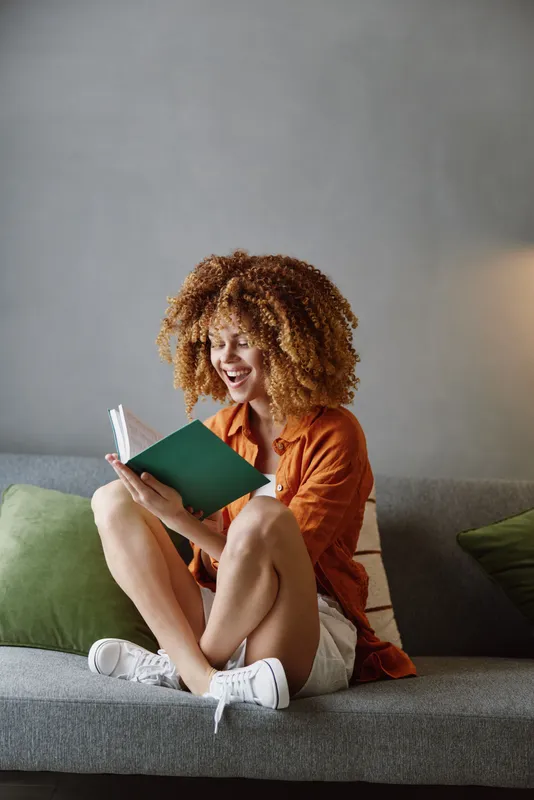 Счастливая женщина читает книгу, сидя на сером диване в окружении зеленых подушек