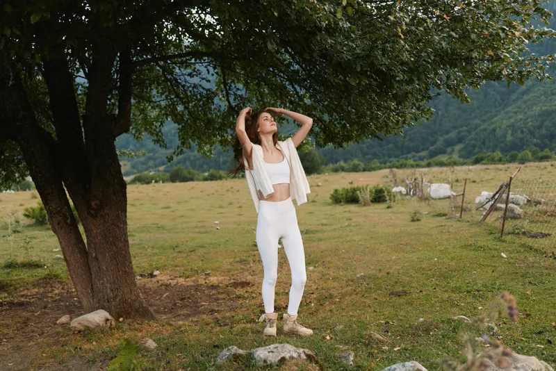 Young woman with long curly hair stands under a tree in a white outfit enjoying nature and fresh air in a peaceful rural landscape.