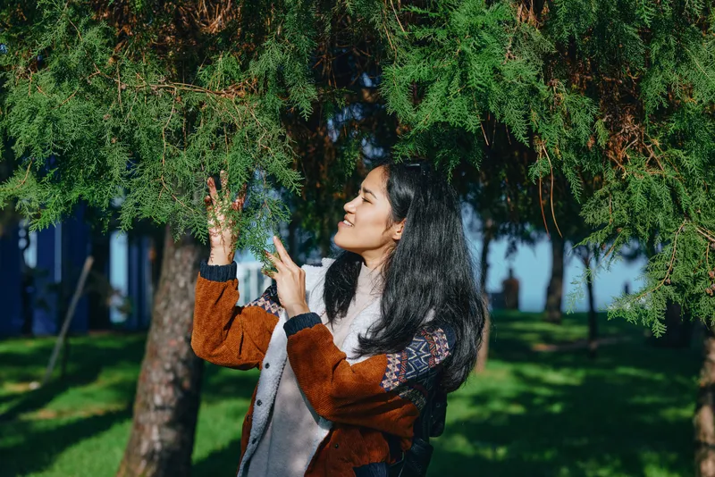 Nature scene with a woman in a warm jacket enjoying light rain under tall trees in a park, reaching toward droplets and smiling, capturing a fresh outdoor mood.