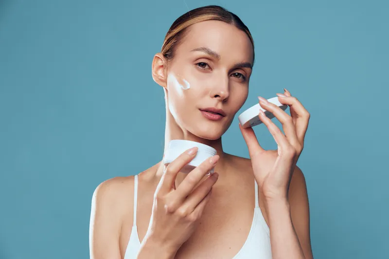Young woman applying face cream with smooth skin, holding jar and cotton pad, skincare routine, clean fresh look, blue background, natural beauty concept
