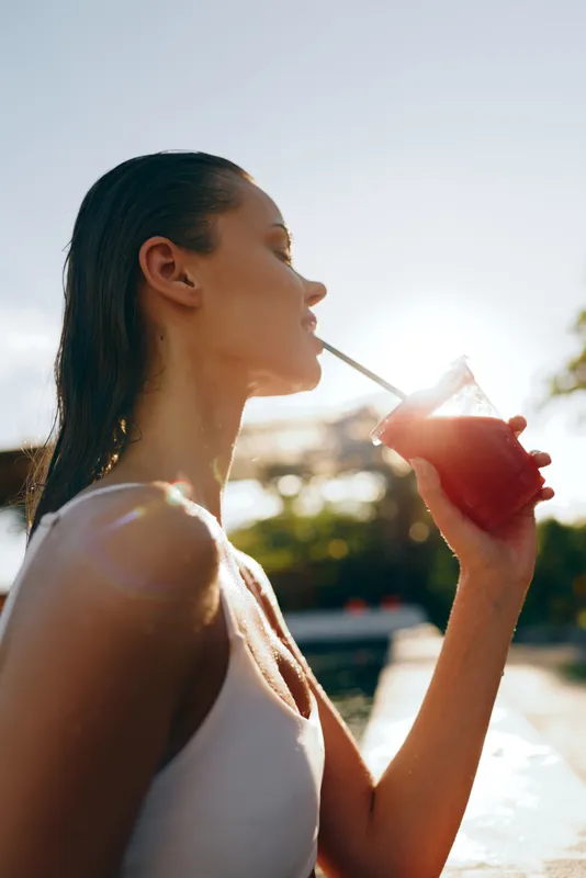 A young woman sips from a straw in bright sun, enjoying a casual outdoor lifestyle moment, relaxed mood, candid expression, warm light, summer vibe outdoors.