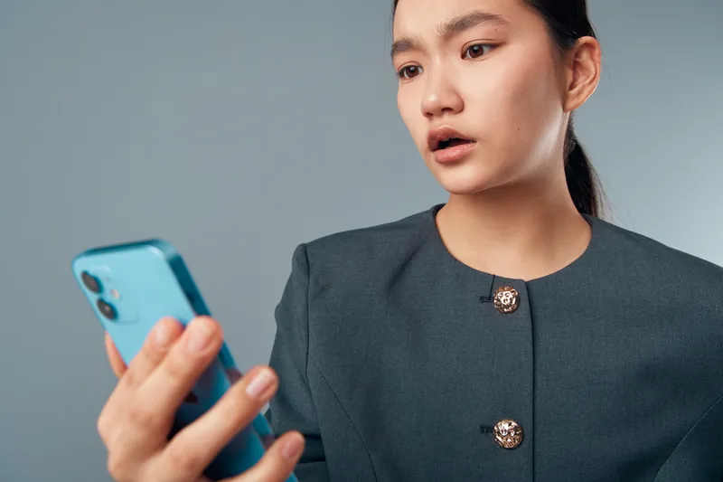 Young woman with dark hair in formal jacket looking surprised at smartphone, holding device with raised hand against gray background in studio. Female, technology and communication concept.