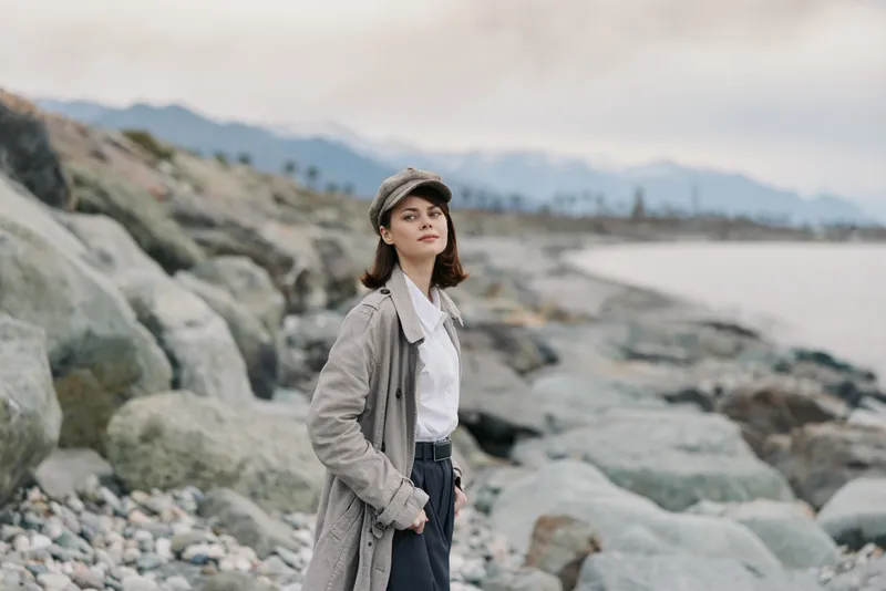 Young woman in casual outfit stands on rocky shore near the sea, looking confidently at camera. Mountains and cloudy sky visible in background, outdoor nature concept.