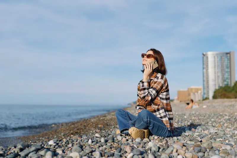 Woman sits on a pebble beach talking on a phone, wearing a plaid shirt, jeans and sunglasses, enjoying sunny outdoor moments by the sea with distant buildings along the shore.