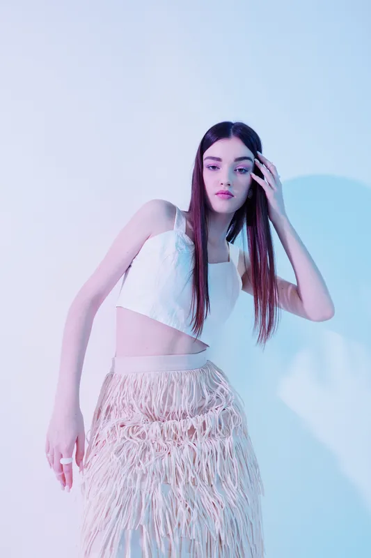 Young woman with long hair poses in a white crop top and fringed skirt against a light background. Concept of beauty, fashion, and style with soft lighting and elegant mood.