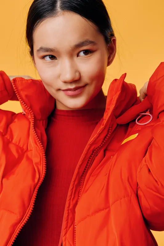 Young woman wearing red winter jacket and sweater, adjusting collar and looking away. Portrait with orange background, casual style, indoors, confident expression and warm clothing for cold weather.