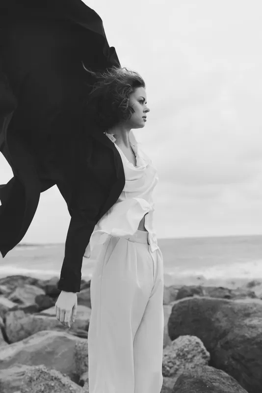 Elegant woman on a windswept beach wearing flowing white pants and a dark coat billowing in the wind, moody monochrome scene, fashion forward, seaside serenity, dramatic silhouette, natural light,