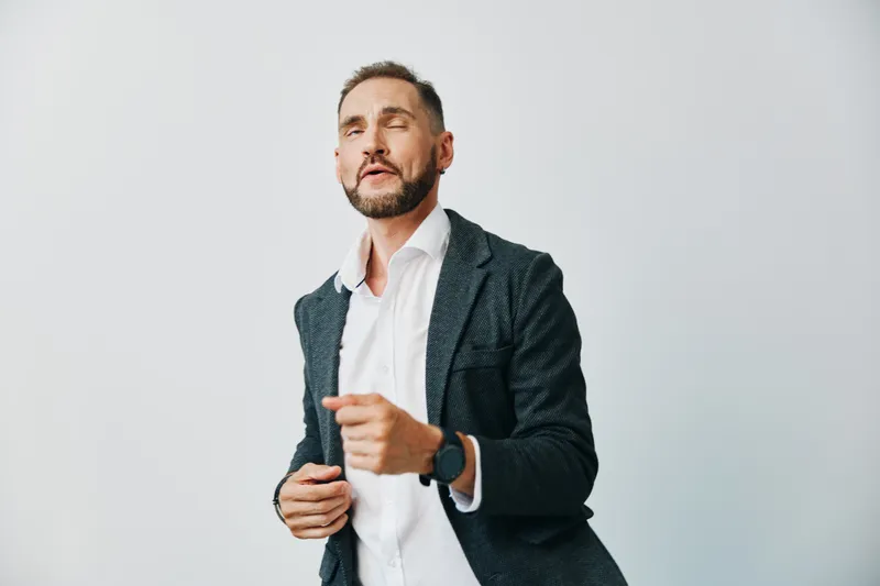 A professional businessman stands against a plain light gray background, projecting confidence. He wears a dark blazer and white shirt, delivering a poised, energetic presence.