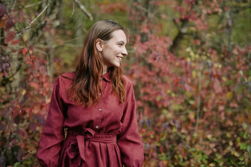 Candid portrait of a young woman smiling among autumn foliage, natural pose and soft light conveying authentic, genuine emotion and grounded presence in an outdoor lifestyle moment.