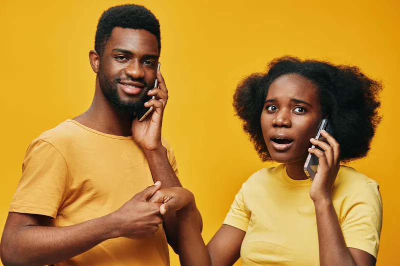 Young African couple communicating excitedly on mobile phones against a vibrant yellow background, showcasing genuine emotions and casual style