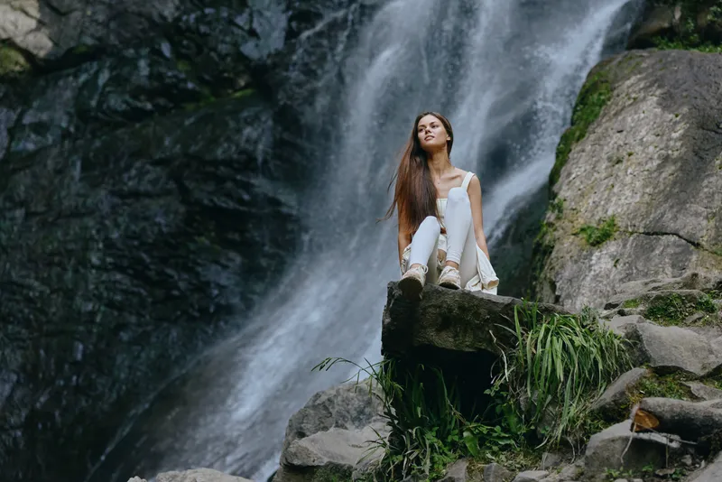 young woman sitting near waterfall in casual white outfit, serene nature setting with rocks and greenery, long hair flowing, peaceful outdoor scene