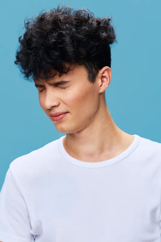 Young man with curly hair wincing and closing eyes tightly against blue background, wearing casual white shirt, expressing discomfort or pain with natural lighting.