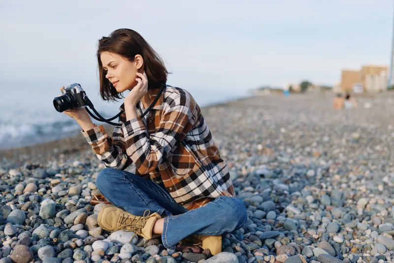 Woman, camera, beach, pebbles, casual, photography: a photographer crouches on a pebble beach in a plaid shirt and boots, concentrating on the ocean horizon and seascape.