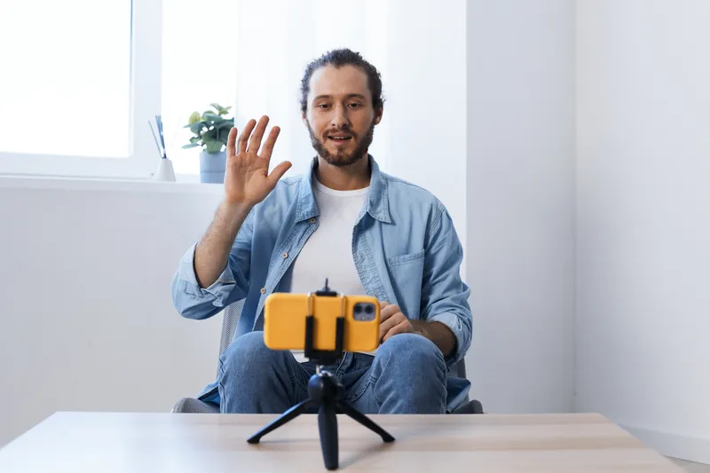 Young man with a casual blue shirt waving during a video call, yellow smartphone on tripod, bright indoor space with natural light, modern concept of remote communication