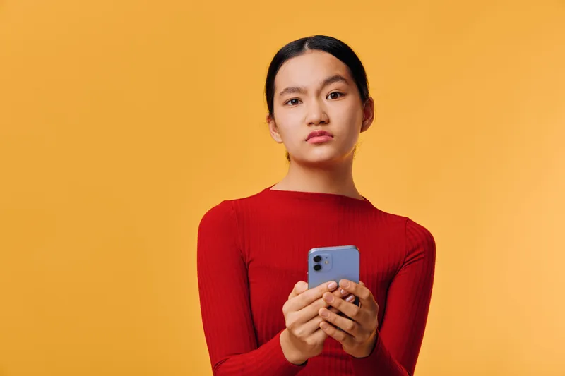 Young woman in red sweater looking serious and holding smartphone with both hands isolated on orange background. Portrait of confident female using mobile device in studio.