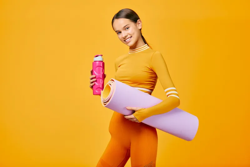 Fitness woman in an orange athletic outfit holds a pink water bottle and rolled yoga mat against a bright orange backdrop, signaling energy, wellness, and sport.