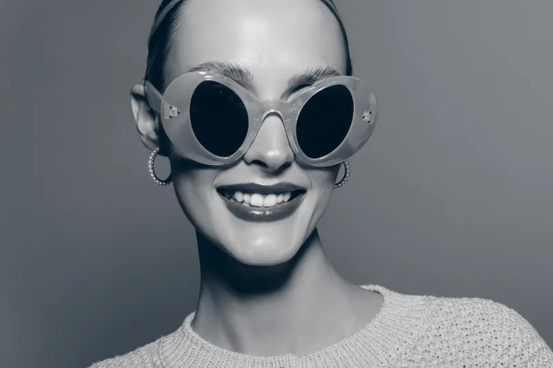 Smiling white woman with pale skin wearing oversized round sunglasses and earrings poses in cozy knit sweater. Fashion portrait with confident expression on neutral studio background.