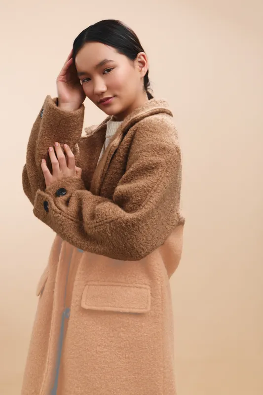 Asian woman wearing a soft beige coat stands in a neutral studio, a gentle smile and poised posture create a calm, stylish portrait with warm lighting and a minimal background.
