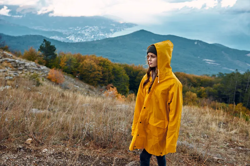 Женщина в жёлтом дождевике стоит на холме, любуясь величественными горами на заднем плане