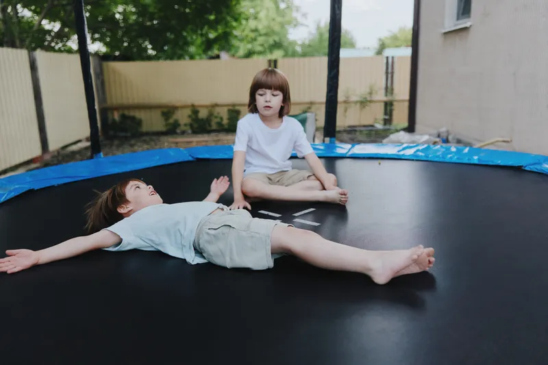 Two children playing on a trampoline, one child lying down with arms outstretched while the other sits. The scene captures joy and carefree childhood moments.
