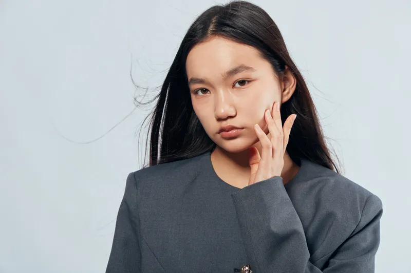 Young woman with long black hair and natural makeup wearing gray sweater posing with hand near face against light background for fashion portrait and beauty concept.