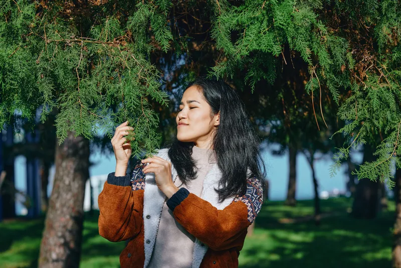 Woman in a warm jacket enjoys sunlit leaves and lush trees in a park, savoring fresh air, calm nature, and a peaceful moment under gentle light