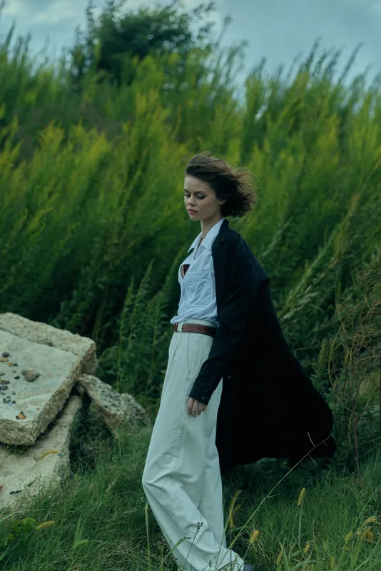 Woman walking in windy outdoor landscape, wearing flowing dress and jacket, amid grass and rocks, with moody sky and distant hills, evoking exploration and calm connection with nature