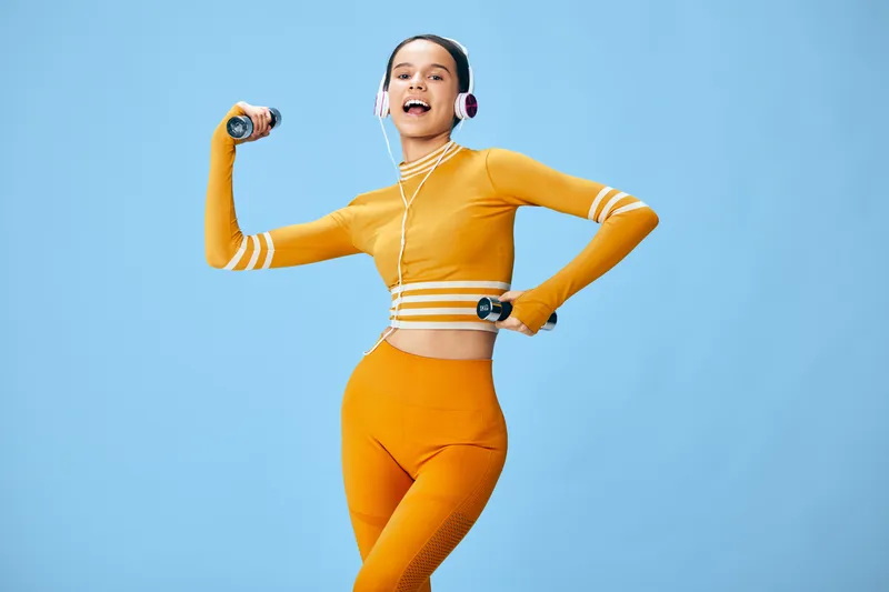 Energetic woman in orange athletic wear poses with dumbbells and headphones against a bright blue background, capturing movement, confidence, fitness passion and a dynamic workout vibe.