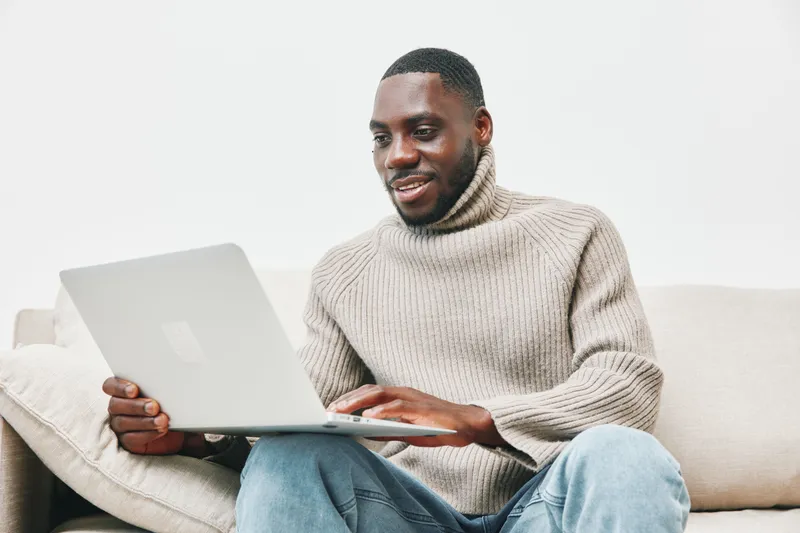 Young African man in a cozy sweater working on a laptop at home, displaying focus and contentment in a soft toned living room setting