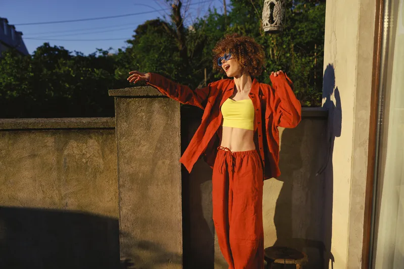 Vibrant woman outdoors, wearing a yellow crop top and red jacket, leaning on a balcony railing, sunlit moment, relaxed pose, warm tones, carefree lifestyle
