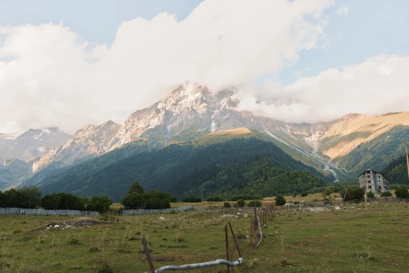 Tranquil mountain landscape with rugged peaks, open pasture, a wooden fence and a distant building under a partly cloudy sky, conveying a peaceful rural outdoor mood today