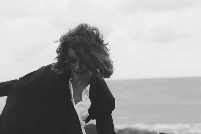 Woman on wind swept beach, coat fluttering, white shirt, black and white composition capturing motion and coastal mood, scene emphasizes energy, texture, and natural light
