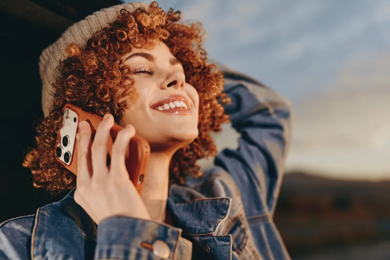 woman with curly hair outdoors, lifestyle portrait, smiling while holding a smartphone near her ear, laughing in daylight, relaxed denim jacket, scenic background, warm breeze