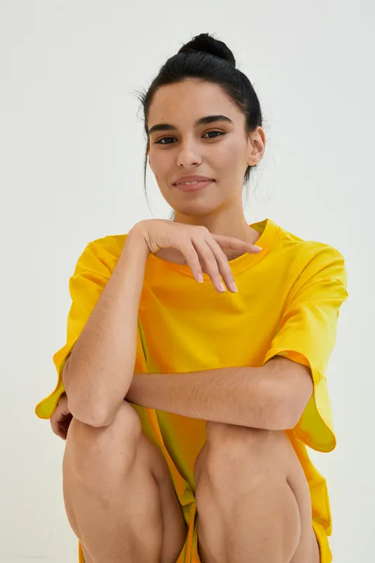 smiling woman in yellow shirt with dark hair tied up sitting casually against white background with natural lighting and relaxed expression