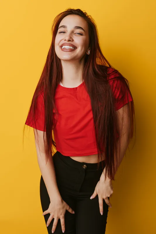 Young woman with long red hair wearing red shirt and black pants smiling with closed eyes standing against yellow background. Casual style and happy female portrait in studio light.