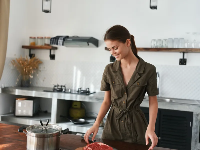 Женщина улыбается на кухне, готовит обед из мяса, овощей и специй на деревянном столе на фоне стильной кухни