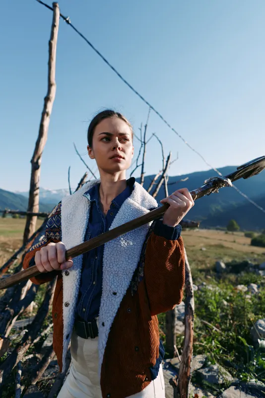 Woman with a wooden staff in countryside mountains, portrait outdoors showing rural traditional style and nature. Young confident traveler by fence in rustic clothing with determined gaze.