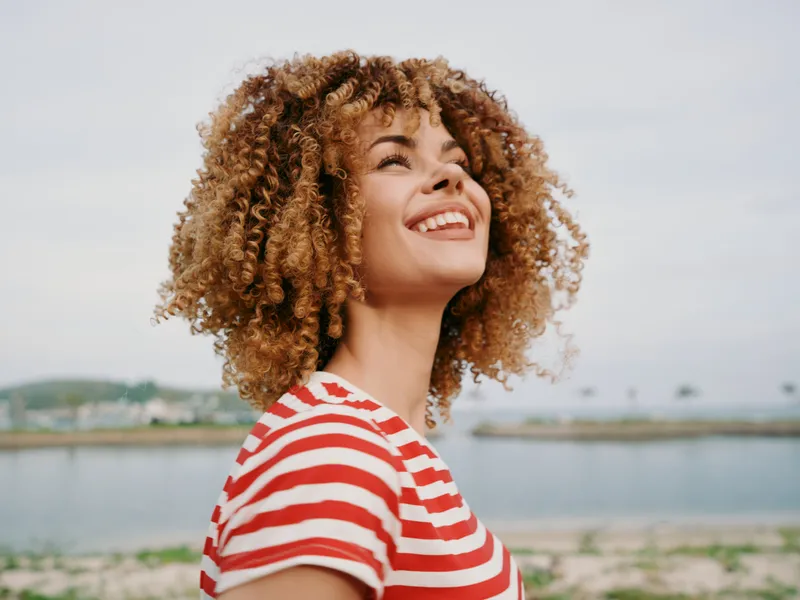 Portrait of a joyful woman with curly hair wearing a red and white striped shirt, smiling toward the sky as sunlight highlights her features near a calm waterfront