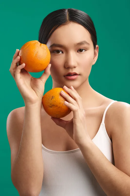 asian woman holds two oranges against a vibrant green backdrop, creating a calm still life with a minimalist, natural mood and soft lighting that emphasizes freshness and balance.
