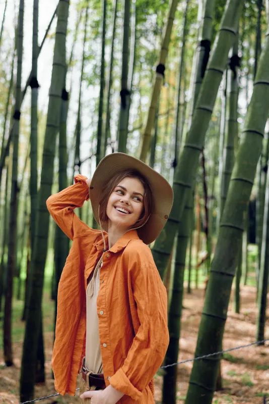 Woman in orange shirt and hat smiling in a bamboo forest, portrait of travel and nature. Young woman enjoying outdoor scenery, relaxed and happy among tall green trees.