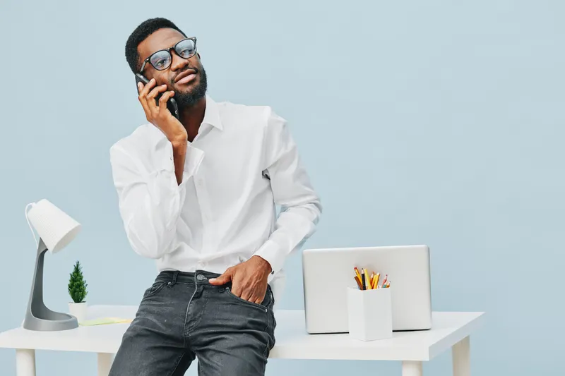 Modern confident man wearing white shirt and sunglasses holding cellphone talking while leaning against desk in bright office workspace with light blue background. People lifestyle concept