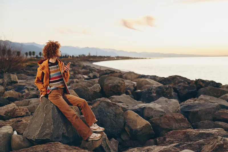 Woman lifestyle scene on rocky seaside shore at sunset, she sits on rocks facing horizon, relaxed and calm, enjoying coastal nature and travel mood, warm sky light.