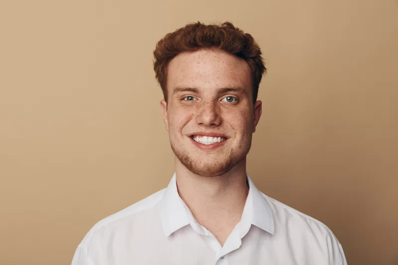 Smiling young man with curly red hair and freckles wears white shirt against beige background. Portrait of confident male model with natural skin and friendly expression in studio lighting.