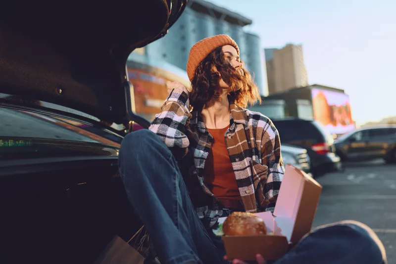 woman car trunk pastry urban golden hour candid lifestyle beanie outdoor - Young woman sits on an open car trunk holding a pastry box in urban parking, warm golden hour glow, authenticity, mindful