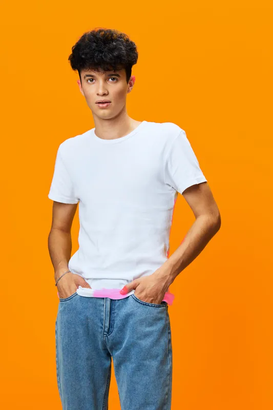 Young man with curly hair wearing a plain white t-shirt and blue jeans stands against a vibrant orange background with a casual and relaxed pose.