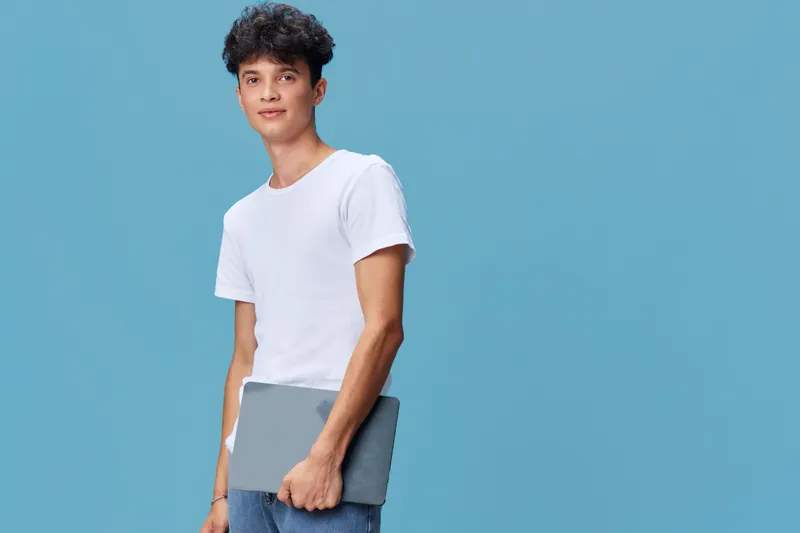 young man holding laptop wearing casual white t-shirt and jeans stands against blue background with relaxed confident expression in bright studio setting