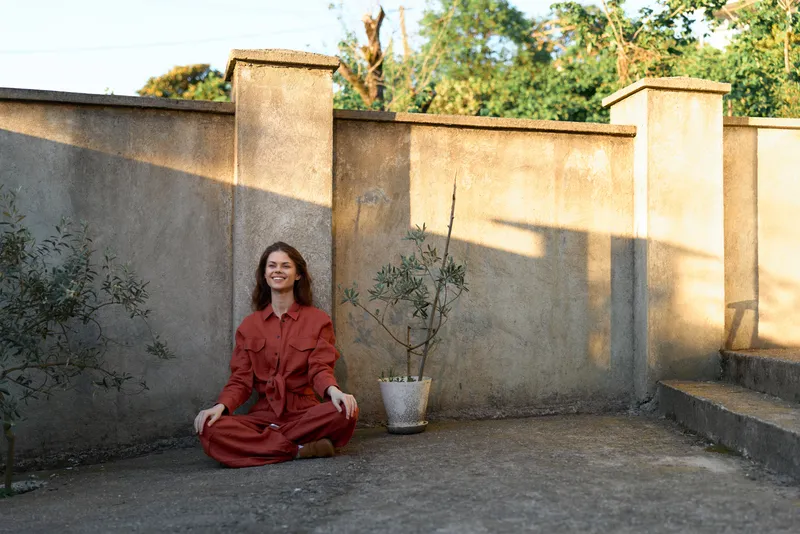 Cheerful woman outdoors, sitting cross legged, red dress, sunlit courtyard, relaxed pose, warm tones, natural textures, serene lifestyle moment in soft light, breeze, earthy backdrop