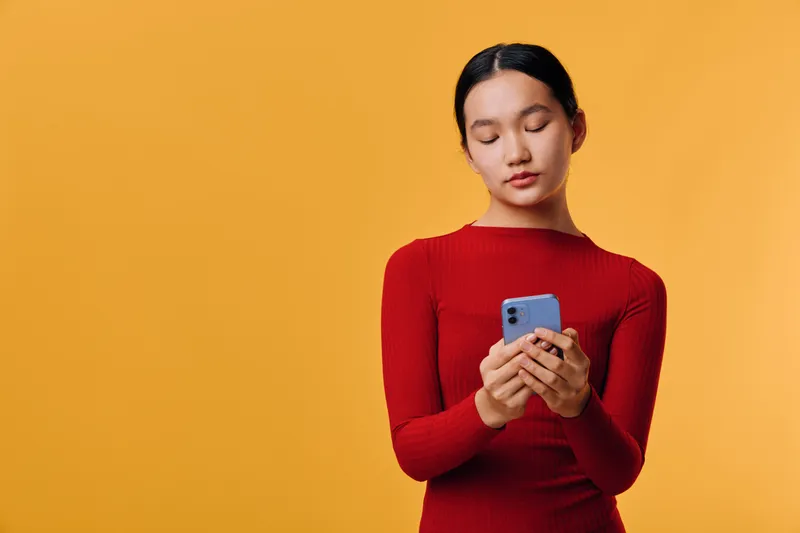 Young woman in red sweater holding and looking at smartphone with neutral expression on yellow background for communication technology and digital connection.