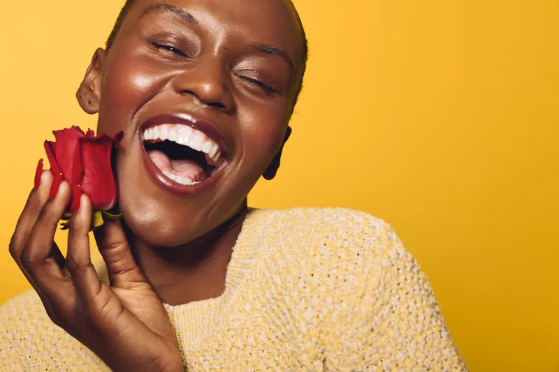 Joyful African woman with dark skin laughing and holding red rose near face. Yellow background and cozy sweater create warm, happy mood. Concept of beauty, happiness, and natural smile.