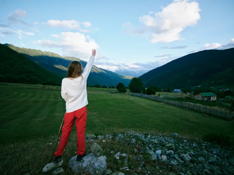 Woman standing on a rocky foreground, raising her hand toward a bright blue sky, with distant mountains and a green valley, conveying exploration, freedom, and outdoor adventure